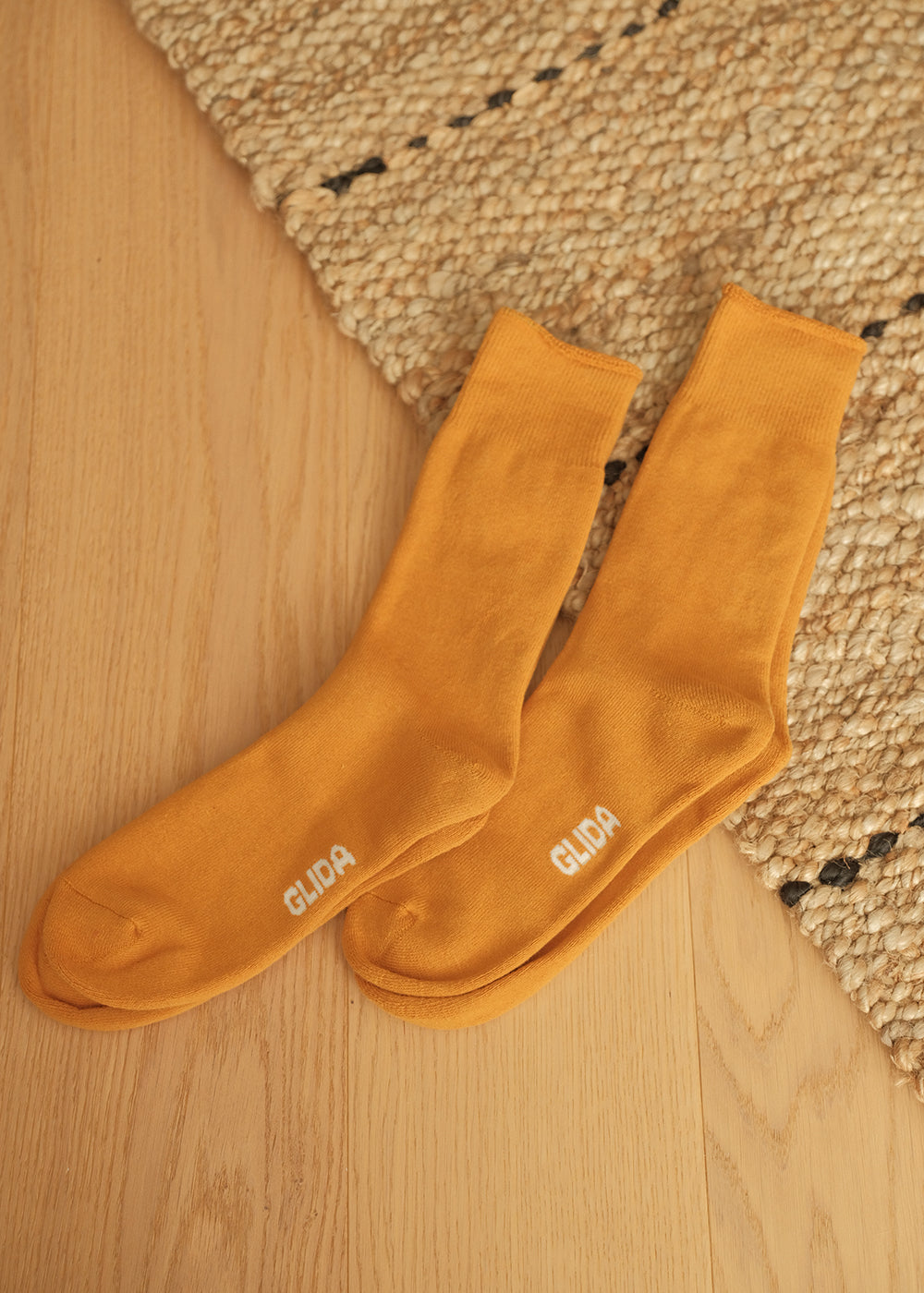 GLIDA Mustard crew socks pair flat lay on wooden floor with woven rug – golden yellow cotton socks with GLIDA branding, overhead lifestyle shot